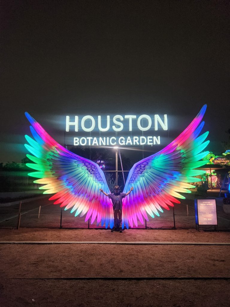 Larger than life rainbow wings with, "Houston Botanic Garden" lit up in white above it. The silhouette of a child is standing in the center of the wings and it is night time so they look like they are the child's wings.